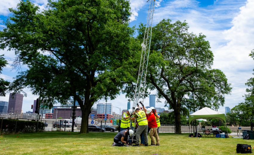 A group of CROCUS team members work on equipment in a sunny patch of grass at UIC. Behind them, tall urban buildings occupy the skyline.